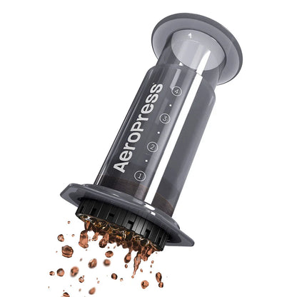 AeroPress con cámara transparente y tapa negra, con café molido saliendo del filtro