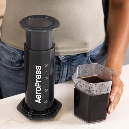 Prensa de café negra con taza de café y mano sosteniendo el vaso