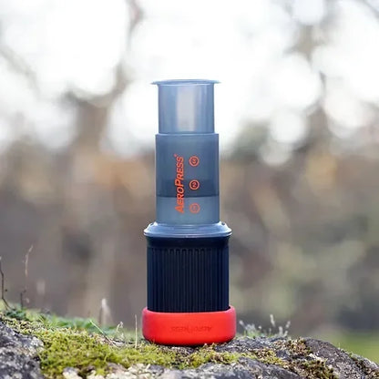 AeroPress GO portátil con base roja y cilindro transparente, apoyado en una roca