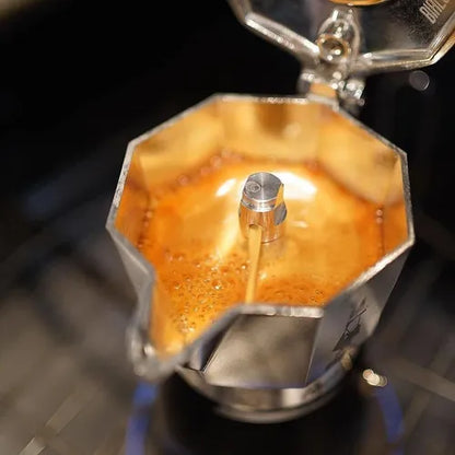 Cafetera italiana con base negra y parte superior octagonal de acero; sale café en preparación.