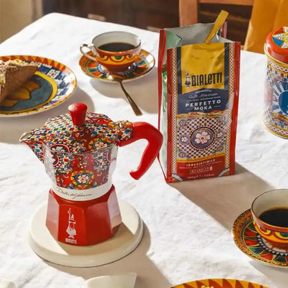 Cafetera moka estampada multicolor con base roja y caja Bialetti, sobre mesa con tazas
