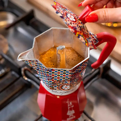 Cafetera moka estampada multicolor vertiendo café, con asa roja y base roja