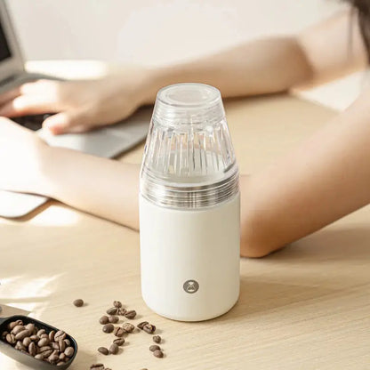 Cafetera portátil blanca con filtro de vidrio cónico, sobre mesa con granos