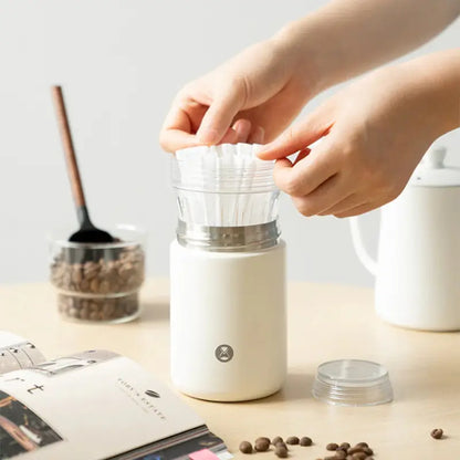 Cafetera portátil blanca con filtro de vidrio abierto; manos colocan el cartucho