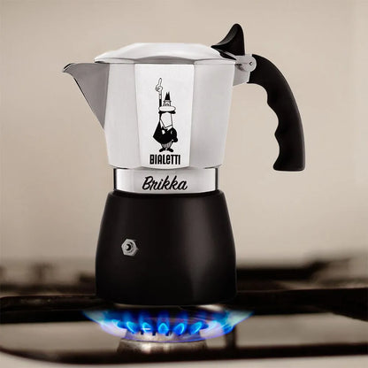 Cafetera italiana de acero con asa negra y base negra, apoyada en una cocina con fuego debajo.