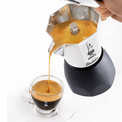 Cafetera italiana de acero octagonal vertiendo café en vaso de vidrio; base negra.