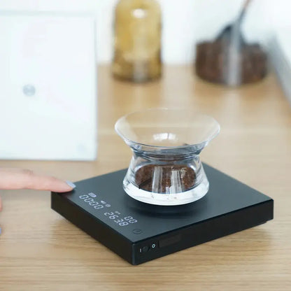 Balanza electrónica negra sobre mesa con taza transparente y lectura digital