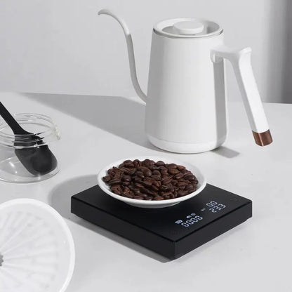 Balanza electrónica negra sobre encimera con taza blanca y recipiente de café
