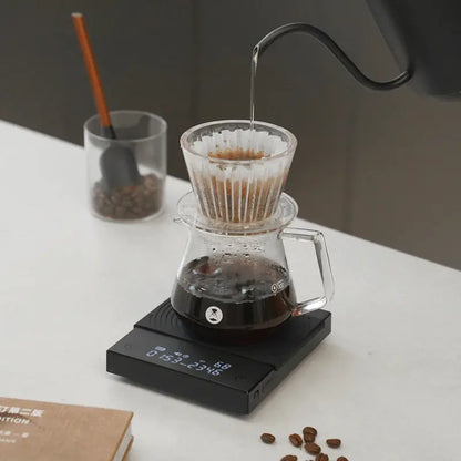 Balanza electrónica negra con cafetera y vaso medidor, lectura en pantalla