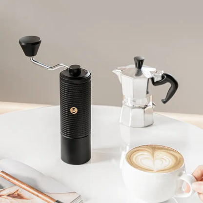 Molino de café manual negro con manivela, junto a taza y cafetera.