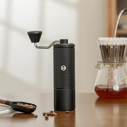 Molino de café manual negro con manivela y base cilíndrica sobre mesa.