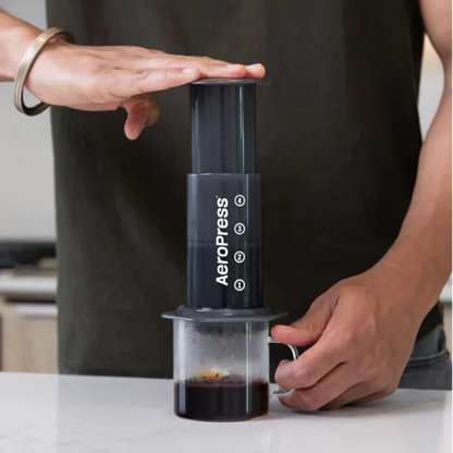 AeroPress en uso con manos, cámara transparente con café y base negra con indicador
