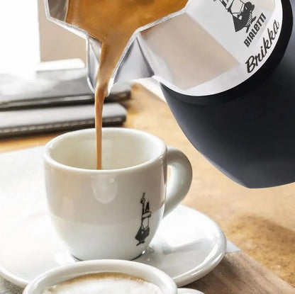 Cafetera italiana con vertido de café desde el depósito octagonal de acero sobre una taza blanca.