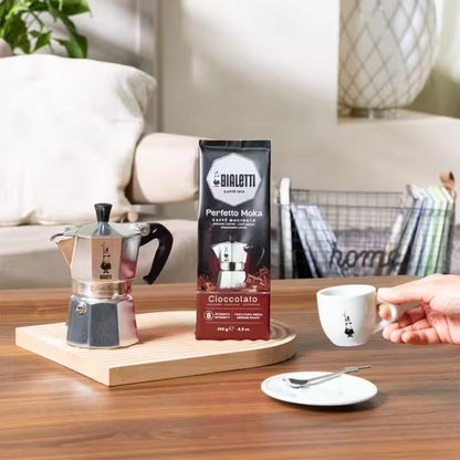 Bolsa de café Bialetti Perfetto Moka Cioccolato, 250 g, junto a cafetera y taza.