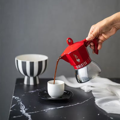Cafetera moka roja Bialetti vertiendo café; jarra con asa y tapa superior.