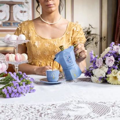Cafetera moka azul celeste con asa dorada y motivo floral, sostenida junto a la taza