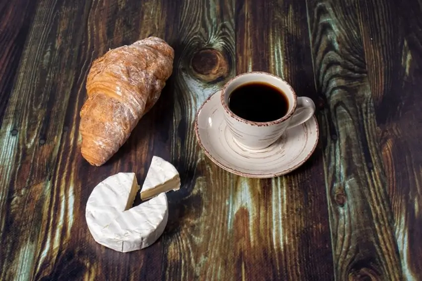 Un croissant crujiente, un queso y una taza de café en una mesa de madera.