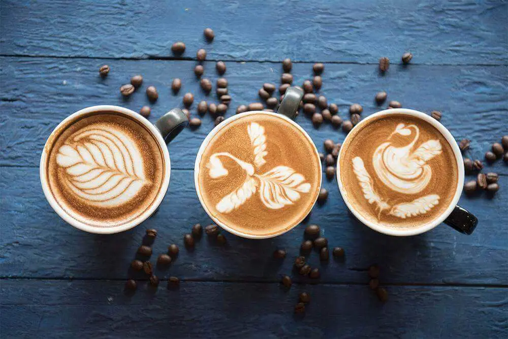 Tres tazas de café con arte latte sobre una mesa azul, mostrando diseños decorativos en la superficie del café.