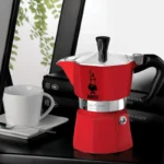 Moka Express roja en un escritorio, acompañada de una taza de café, ideal para disfrutar de un espresso italiano.
