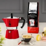 Cafetera Moka Express roja sobre una mesa con una bolsa de café y una taza, diseñada para preparar 3 tazas 130ml de espresso italiano.