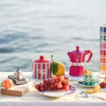 Mesa con frutas frescas, café en cafetera italiana Bialetti y otros elementos decorativos, evocando un ambiente veraniego.