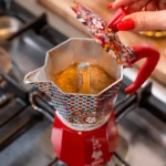 Persona abriendo la tapa de una cafetera italiana Bialetti roja, decorada con motivos de carretos sicilianos de Dolce&Gabbana.