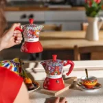 Mujer con una cafetera roja Bialetti en la cocina, un diseño que celebra la tradición siciliana y el arte italiano.