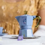 Cafetera Moka Italiana sobre una mesa con una taza de té, decorada con flores y abejas, inspirada en Bridgerton.