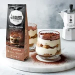 Café en vaso con cuchara y cafetera, fusionando el sabor del tiramisú italiano en un espresso rico y cremoso.